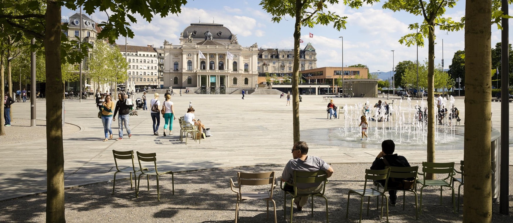 Das Bild zeigt den Sechseläutenplatz an einem heissen und sonnigen Tag. Menschen sitzen auf Stühlen unter Bäumen und Kinder spielen im Wasserspiel. 