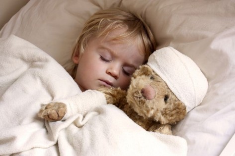 Ein kleines Kind liegt krank im Bett, sein Teddybär trägt einen Kopfverband