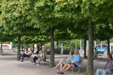 Menschen sitszen auf Bänken unter den Bäumen. 