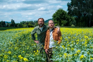 Das Bild zeigt die beiden männlichen Gründer der Firma inmitten einem gelbblühenden Feld. 