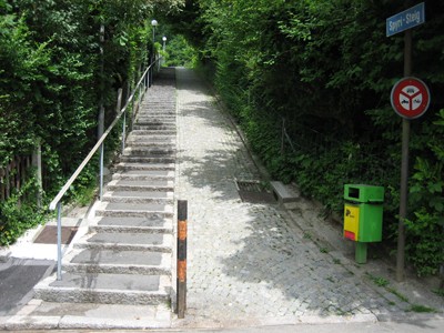 Spyri-Steig. Treppenweg gepflästert, mit Handlauf und Schieberampe, abgegrenzt durch Sträucher und Hecken.